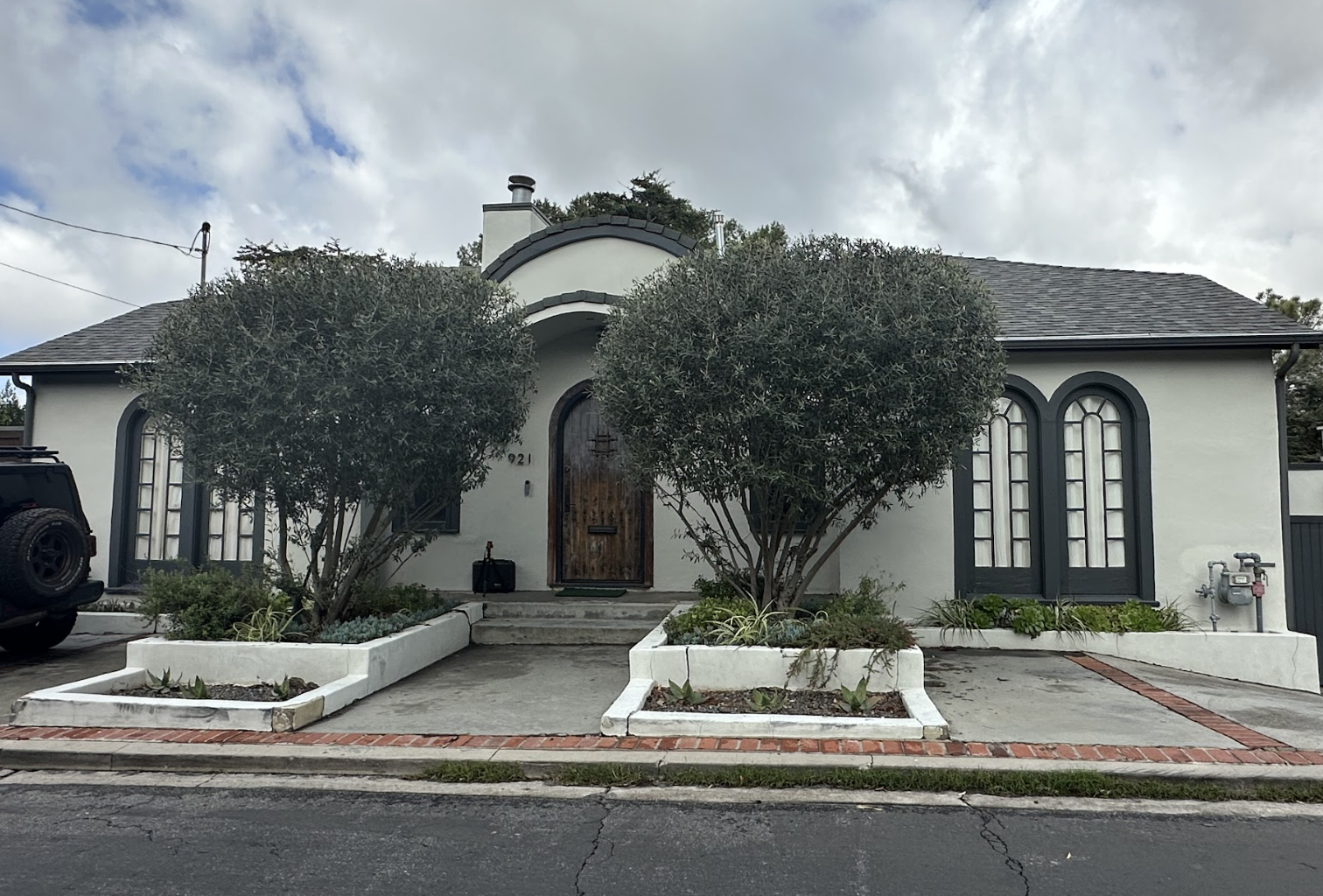 User-Inserted A Charming White Stucco House With Arched Windows And A Wooden Door, Framed By Two Manicured Trees In Rectangular Planters On A Cloudy Day.