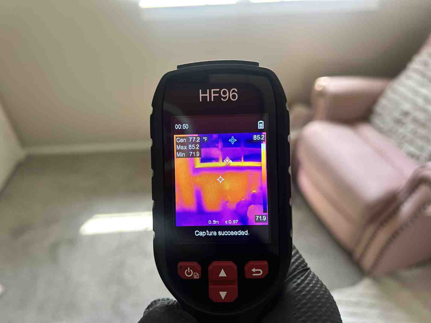 User-Inserted Close-Up Of A Handheld Thermal Imaging Device Displaying A Heat Map Of A Room, Showing Temperature Variations In Pink, Purple, And Orange Tones.